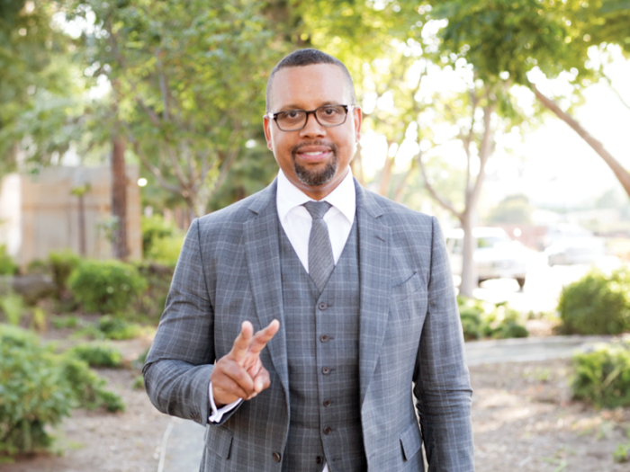 Shay Dean is wearing a gray suit. He's standing outside, surrounded by greenery. He's smiling at the camera and holding up his right hand in a peace sign.