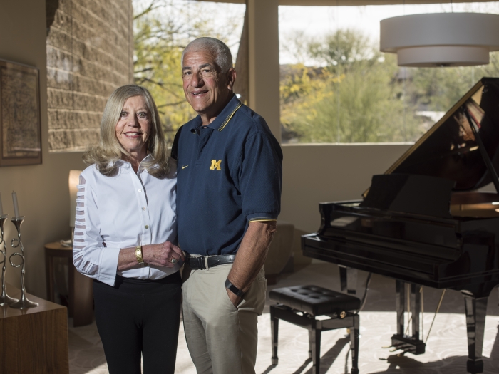 Susan and Richard Rogel pose next to a grand piano. Rogel is wearing a polo shirt with a block M.